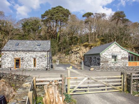 Two stone buildings with slate roofs behind a wooden gate and a gravel driveway with trees on a hill behind at Tyn Llwyn Cornel Eco Barn in Garreg