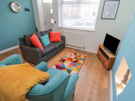 A living room with a gray sofa and colorful cushions a teal armchair with a yellow throw a small wooden TV stand with a television and a multicolored rug on the floor at Sea Glass in Filey