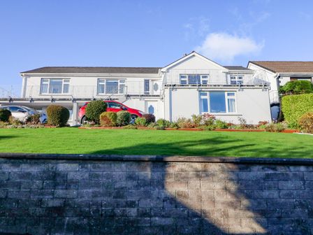 A white two-story house with a balcony driveway and parked cars behind a stone retaining wall and lawn at Lavender Cottage in Castletownbere County Cork
