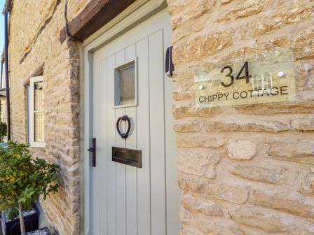 A pale green front door with a heart-shaped knocker and a letterbox on a stone wall with a house number 34 sign at Chippy Cottage in Chipping Norton
