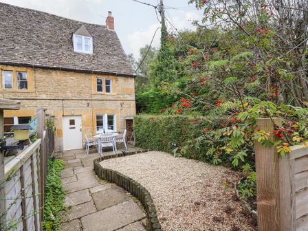 A stone cottage with a tiled roof and white door next to a patio with outdoor furniture and a gravel area with plants at Gleed Cottage in Naunton