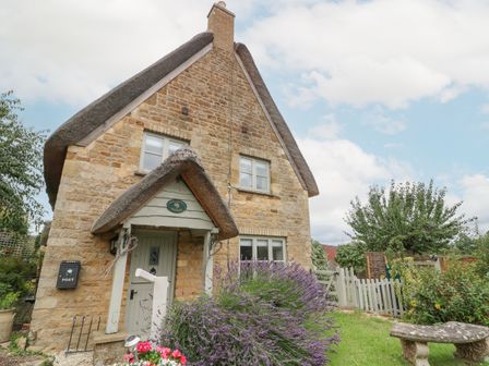 A stone cottage with a thatched roof surrounded by a garden with flowers and a bench at Honeysuckle Cottage Brailes