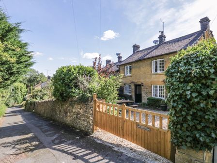 A stone cottage with windows and chimneys behind a wooden gate and stone wall along a driveway at Jackdaw Cottage in Blockley