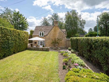 A garden with a lawn, flower beds, and hedges in front of a stone cottage with an umbrella at Weir Cottage in Bourton-on-the-Water