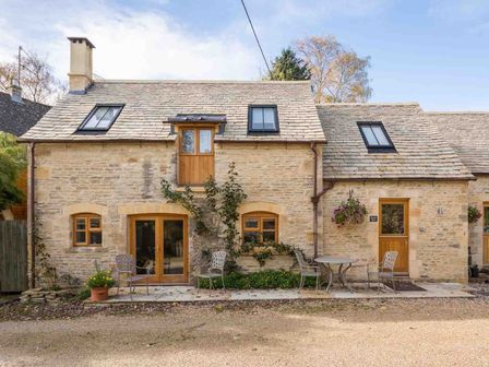 A stone cottage with wooden doors and windows and outdoor chairs and table at Oyster Barn in Naunton