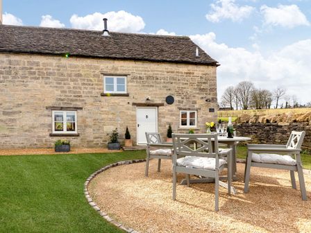 An outdoor seating area with a table and four chairs on gravel in front of a stone building with windows and a door at The Creamery in Nympsfield