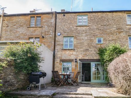 A stone patio with a wooden table and chairs and a barbecue outside a stone house at Red Fawn Cottage in Blockley