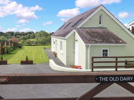 A light green house with a paved driveway a grassy yard and wooden gate labeled The Old Dairy in Maenclochog