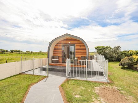 An arched wooden cabin with two chairs on the porch and a fenced patio in a grassy area at Caban Cariad in Bryngwran near Bodedern