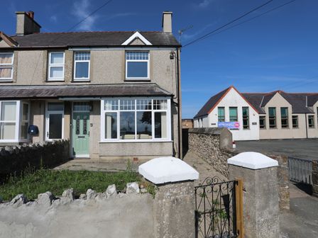 A semi detached house with a green door and front garden next to a stone wall and a building with multiple windows at The Old School House in Bodorgan