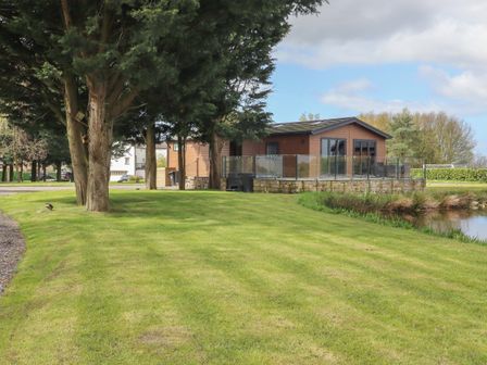A wooden house with a glass railing on a stone base near a body of water and trees on a grassy lawn at Ruperts Retreat in Nateby near Garstang
