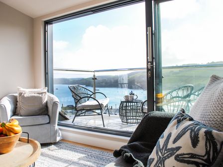A living room with a cushioned chair fruit bowl and pillows facing a balcony with chairs and a table overlooking a body of water at Higher Cliftonville in Salcombe