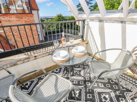 A balcony with a glass table set for two and two chairs overlooking trees and buildings at Puffins in Tenby