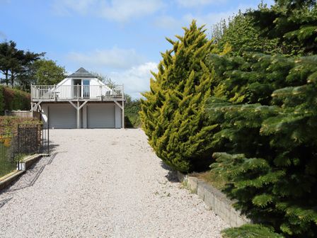 A gravel driveway leading to a two garage doors with a balcony and white structure above surrounded by trees at Higher Gitcombe Lodge in Dartmouth