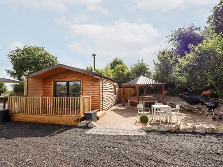 A wooden cabin with a deck and sliding glass door next to a pavilion with a hot tub and outdoor table with chairs surrounded by trees at Celyn in Cyffylliog