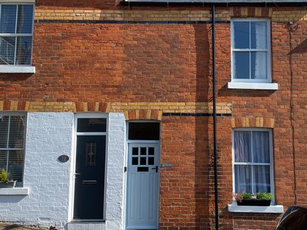A brick house exterior with two doors and four windows one door and window have white paint around them and there are flower boxes under two windows