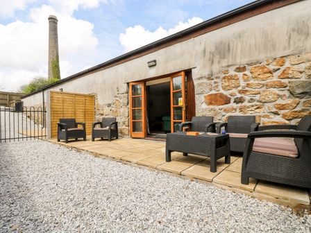An outdoor patio area with black wicker chairs and a table on stone tiles outside a stone building at Wheal Prosper@The West Dry in St Austell