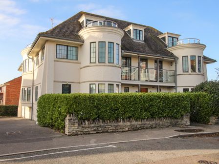 A white two-story house with curved bay windows and a glass balcony behind a hedge at Broadleys in Swanage