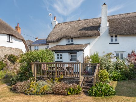 An outdoor wooden deck with chairs and a table surrounded by plants and a thatched roof house at Rose Thatch in Malborough