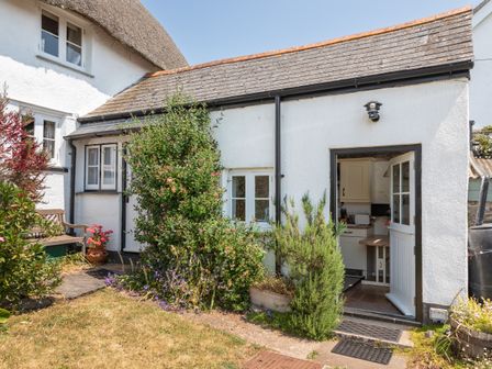 The exterior side view of a white cottage with an open door showing part of a kitchen and various plants and bushes outside the cottage at End Cottage in Malborough