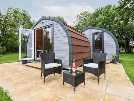 Two small pod-shaped cabins on a patio with two chairs and a table holding a bottle and two glasses at Meadow Lodge Berrys Place Farm in Churcham
