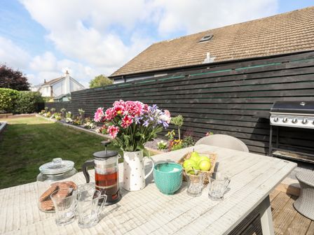 An outdoor patio table with flowers fruit mugs and a teapot near a black fence and a barbecue grill at Dolwyn in Llandegfan