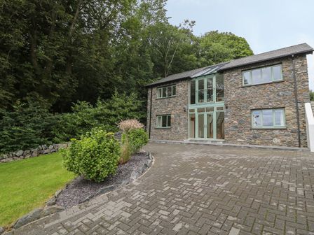 A stone house with green window frames and a paved driveway next to bushes and trees at Tan Twr in Llanfairpwll