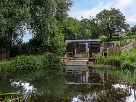 A small cabin with outdoor seating overlooking a pond surrounded by trees and greenery at The Shepherds Hut at Bridge Lake Farm & Fishery in Chacombe