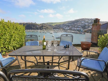 An outdoor patio with metal furniture and blue striped cushions overlooking a harbor with boats and hills at Juniper Cottage in Dartmouth