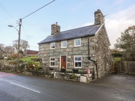 A stone cottage with a wooden door and four windows by a road with a wooden gate and greenery at Ty'n y Clawdd in Dyffryn Ardudwy