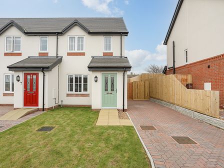 A front view of two houses with a red door and a green door with a lawn and paved driveway at Swn Y Nant in Holyhead