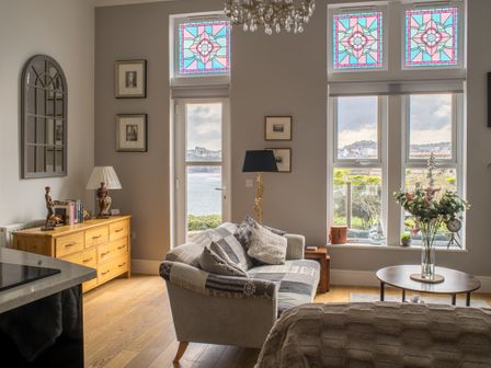 A living room with a sofa, wooden chest of drawers, coffee table with flowers, and windows with stained glass at King Edwards View in Newquay