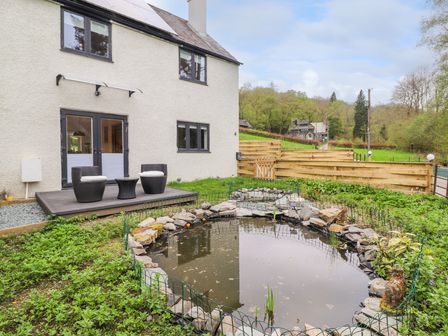 A garden with a pond surrounded by rocks and a padded seating area outside a two-story house at Yamthwaite in Grizedale near Hawkshead