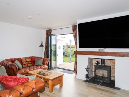 A living room with leather sofas pillows a wooden coffee table a floor lamp a wood stove under a large wall mounted television and bi fold doors leading outside at The Hawthorns in Llanfechell