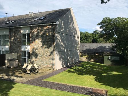 A stone and plaster house with a patio set on a paved area next to a lawn and a green shed in the garden at Barn End 9 Stad Clynnog in Newborough