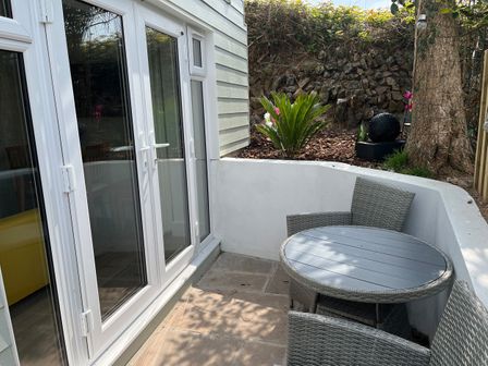A small outdoor patio with a round table and wicker chairs next to white double doors and a garden area with plants at Perran Pearl B in Perranporth