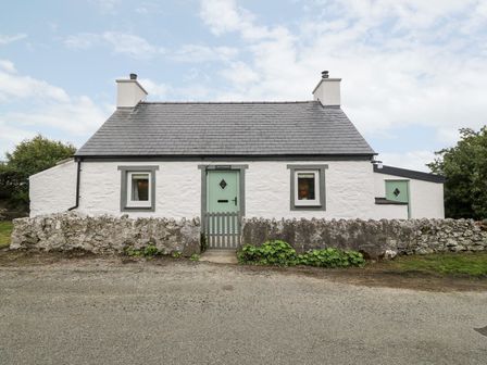 A white stone cottage with two chimneys two windows and a green door behind a low stone wall at Rhytty Bach in Ty Croes near Rhosneigr