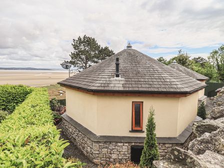 An octagonal stone and stucco building with a shingle roof near a beach with hedges and trees at Oversands Cottage in Grange-Over-Sands