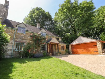 A stone house with green window frames a wooden front door a gravel driveway and a wooden garage door surrounded by grass and trees at 1 The Lane in Chipping Norton