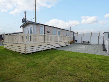 A mobile home with a surrounding deck and fenced gravel area with picnic table and barbecue on grass at Trewan Lodge in Bryngwran near Rhosneigr