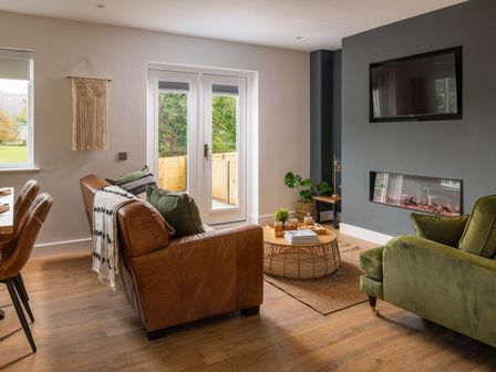 A living room with a brown leather sofa a green armchair a round wicker coffee table a wall-mounted fireplace and a television at Sygun in Beddgelert