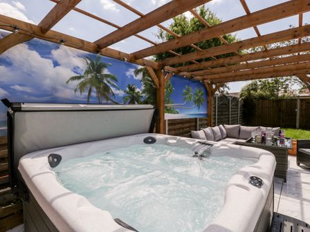 An outdoor area with a hot tub under a wooden pergola next to a seating area with a table and chairs and a tropical mural on the wall at Seacider in Burnham-On-Sea