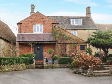 A brick house with a black door and statues of dogs by the steps surrounded by shrubs at The Cheese Room in Stretton-On-Fosse