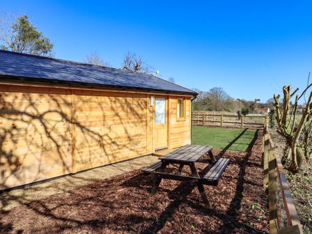 An outdoor area with a wooden picnic table beside a wooden building and a fenced grassy area at Buttercup Lodge in Theberton near Leiston