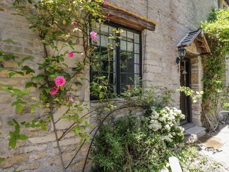 A stone cottage wall with climbing plants and flowers around a window and door at Fox Den Cottage in Castle Combe