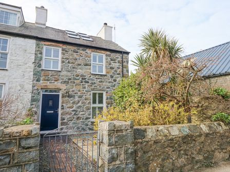 A stone house with a blue door behind a metal gate and stone wall with plants and trees at Min Y Mor - Morfa Nefyn