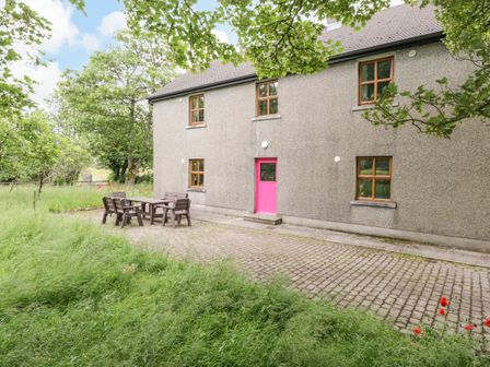 A gray two-story house with wooden framed windows and a pink door with outdoor wooden table and chairs on a paved area surrounded by grass and trees at Bohans Barn in Crossmolina County Mayo