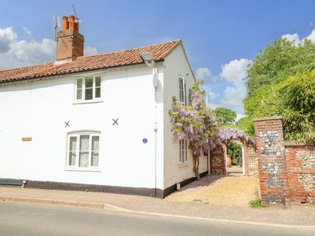 A white two-story house with a tiled roof and chimney next to a brick and stone archway with purple flowering plants at 1 Chelsea Cottage in North Elmham