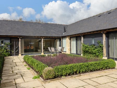 An outdoor courtyard with a square garden bed surrounded by hedges and patio furniture at Hawthorn Barn in Northleach