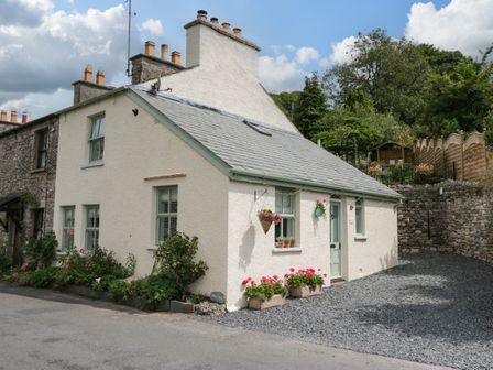 A cream cottage with green window frames and flower pots outside on a gravel driveway at West View in Leasgill near Milnthorpe
